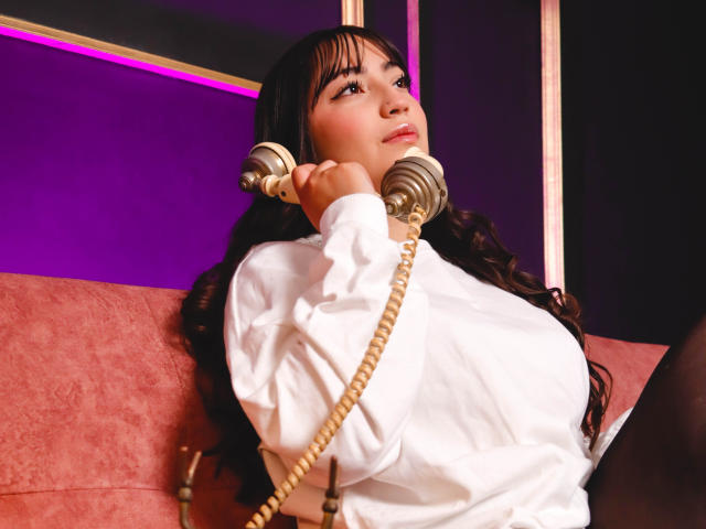 Ann Fisher, avec ses longs cheveux noirs, portant une chemise blanche, tenant un vieux téléphone à l'oreille, regardant vers le haut avec un léger sourire, assise sur un canapé rose..
