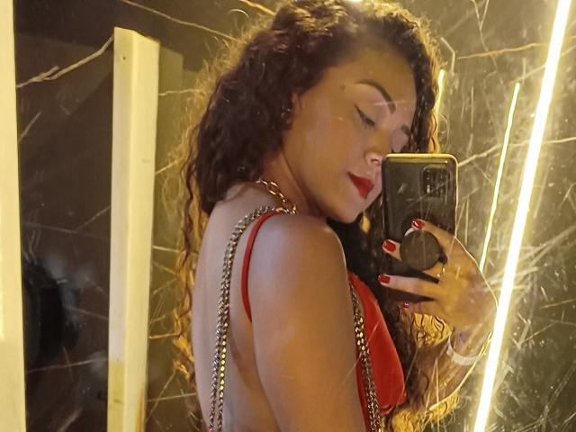 LaisMiguel with curly red hair, red lipstick, and red bra, taking a mirror selfie, showing her bare back and sideboob, with a chain necklace and red nails.