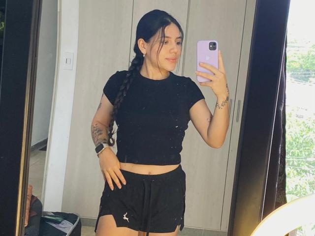 SophiaMarx with long black braid, black crop top, and black shorts, holding a purple phone, showing tattoos on her left arm, smiling, standing in front of a mirror.