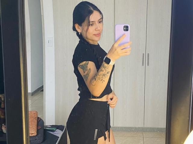 SophiaMarx with tattoos on her arm, black top, and black shorts, holding a purple phone, taking a mirror selfie, showing her ass and a wristwatch.