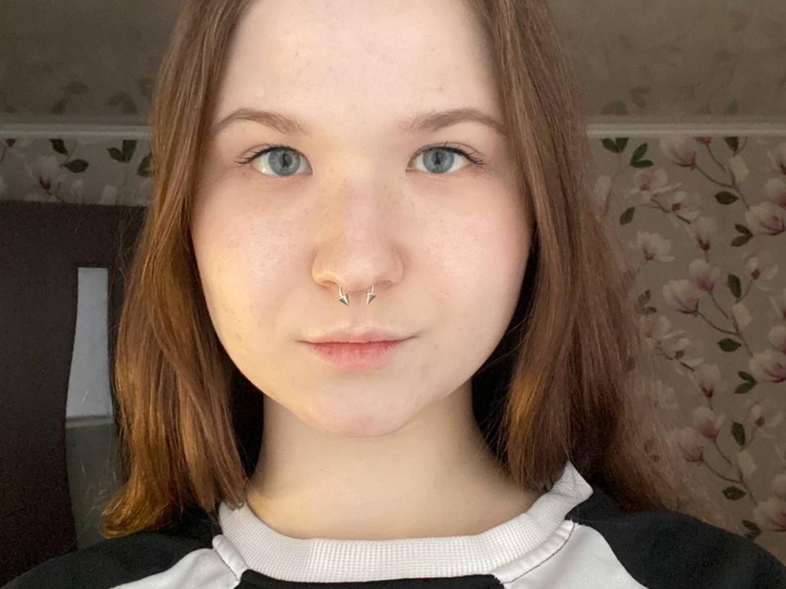 Pam Gillilan tem pele clara, olhos azuis, cabelo castanho e usa um piercing no nariz. Ela está vestindo uma camisa preta e branca com uma gola rendada. Suas nádegas não estão visíveis..
