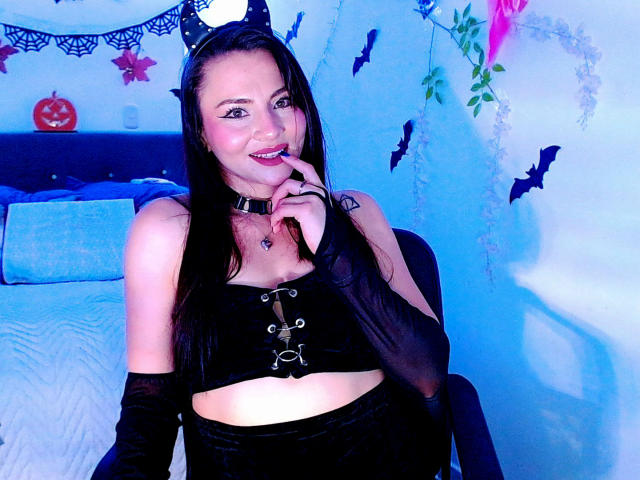 EmelySofia con orecchie da gatto nere, indossa una crop top nera con lacci, maniche nere e un collare, sorride con un dito sulle labbra, mostrando il seno e un piccolo tatuaggio..