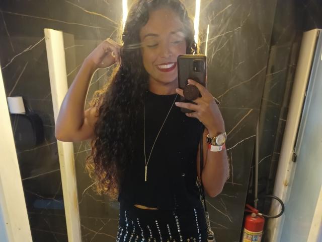 LaisMiguel with curly brown hair, smiling, wearing a black sleeveless top, necklace, and studded black pants, holding a phone in a bathroom mirror selfie.