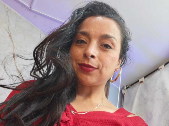 StangelarRivera with wavy black hair, red lipstick, and red shirt, wearing hoop earrings and necklace, smiling slightly, showing small mole on left cheek.