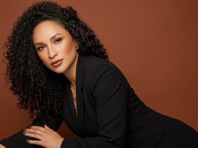 CarlaMuille with curly black hair, wearing a black blazer, looking at the camera with a serious expression, brown eyes, and subtle makeup, hands resting on a surface.
