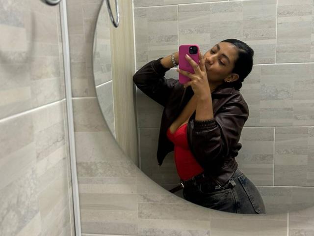 NatalyBlonder mit dunkler Haut, schwarzen Haaren im Dutt, trägt ein rotes Oberteil, eine braune Jacke und blaue Jeans und macht ein Selfie vor einem Spiegel mit einem rosa Handy. Brüste sind sichtbar..