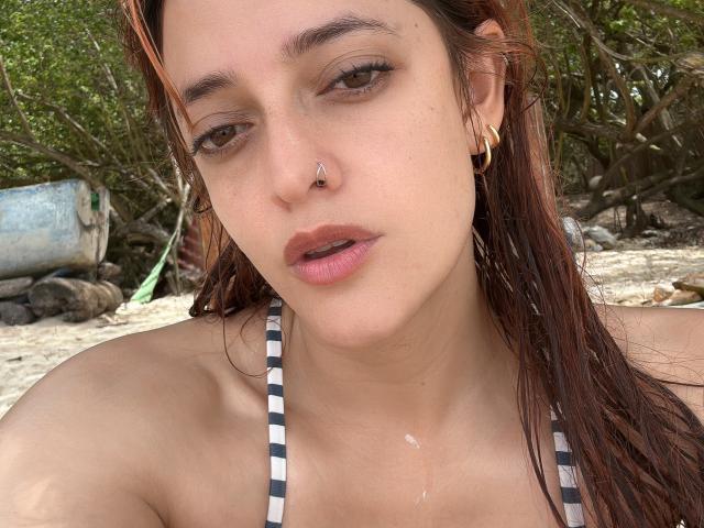 JaneRoa, con i capelli castani bagnati, un piercing al naso, orecchini dorati e un top da bikini a righe, mostra leggermente le labbra rosa e una piccola goccia d'acqua sul petto..