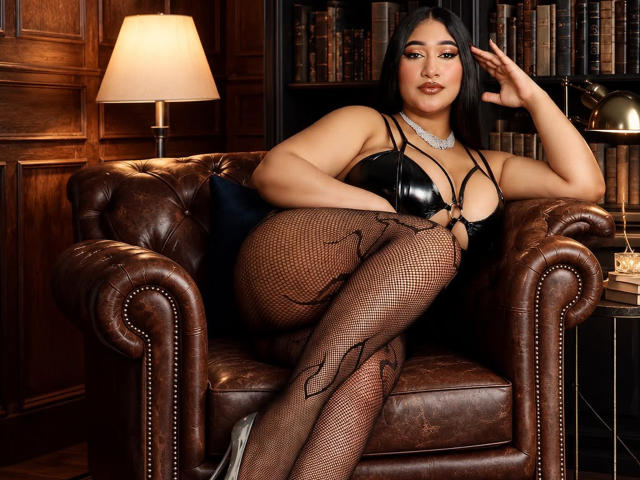 MelisaJackie with big boobs, wearing a black leather bra, fishnet stockings, and heels, lounging on a brown leather chair, looking sexy, dark hair, silver necklace, confident expression.