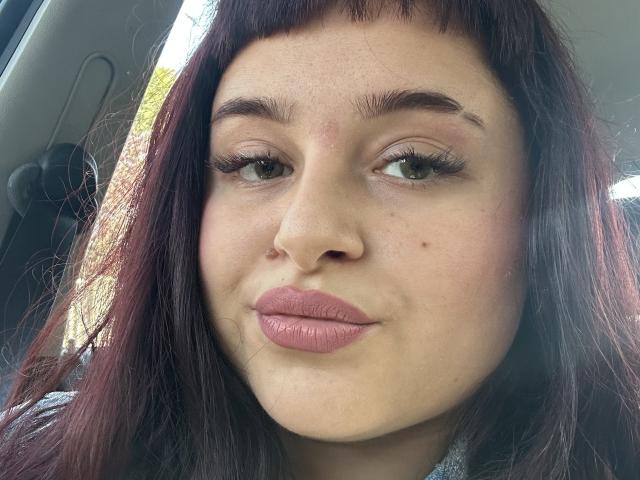 Luucie has a cute face with brown eyes, dark red hair, and pinkish lips, showing a slight smile. She's got a small zit on her forehead and a beauty mark.
