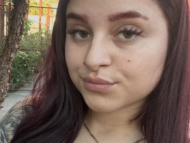 Luucie has dark red eyebrows, long eyelashes, and pinkish lips with a slight smirk. She has light skin, a small mole on her right cheek, and dark brown hair.