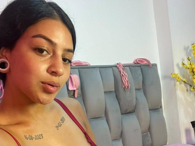 LonxEva with dark hair, brown eyes, and pink ear gauges, wearing a red top, showing her chest tattoo, and a subtle pout. Her tits are small to medium.