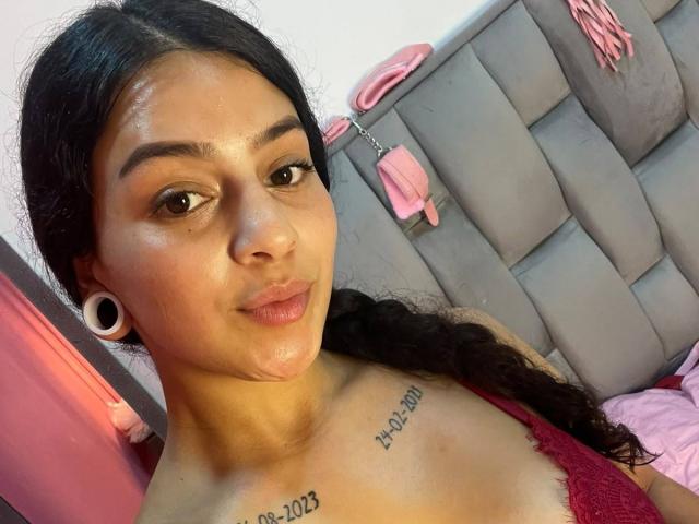 LonxEva with shiny skin, dark eyebrows, and long black braids, wearing a red lace top, showing off large white ear gauges and a tattoo above her tits.