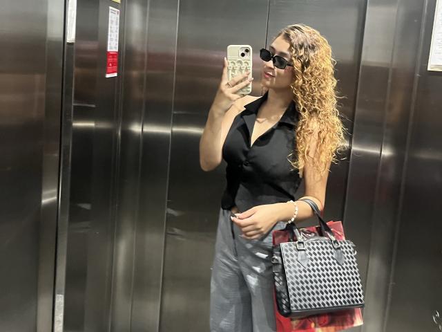 Arianna Blake, avec ses cheveux blonds bouclés, son t-shirt noir, son pantalon gris, ses lunettes de soleil, tenant un téléphone, un sac à main noir à motifs et un sac rouge, se tient dans un ascenseur en métal, exhibant ses fesses volumineuses..