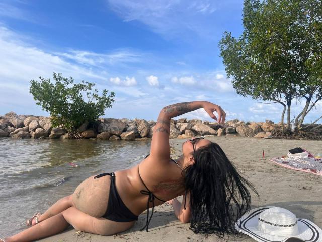AlanaLuxe allongée sur une plage de sable, en maillot de bain noir, avec un bras tatoué, de longs cheveux noirs, les fesses en l'air, un chapeau de soleil sur la droite, un ciel bleu, des rochers, de l'eau, des arbres, et un marqueur rouge en arrière-plan..