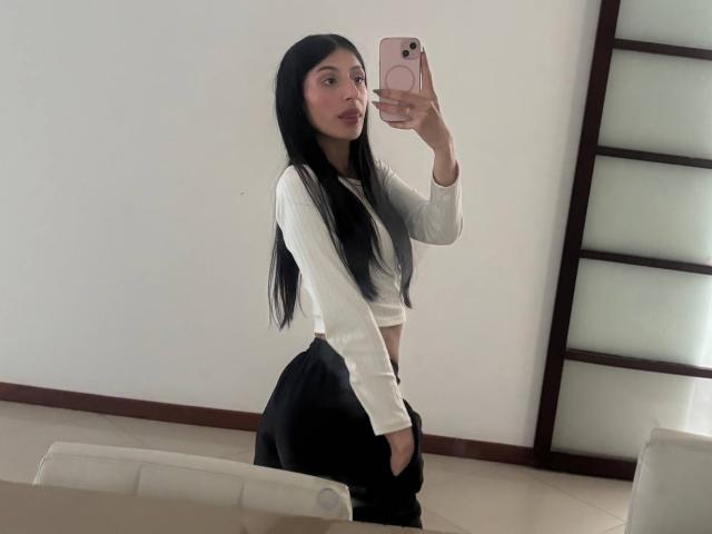 SophieBeaumont with long black hair, white ribbed top, black pants, taking a mirror selfie, showing her ass, holding a pink phone, in a minimalistic room.