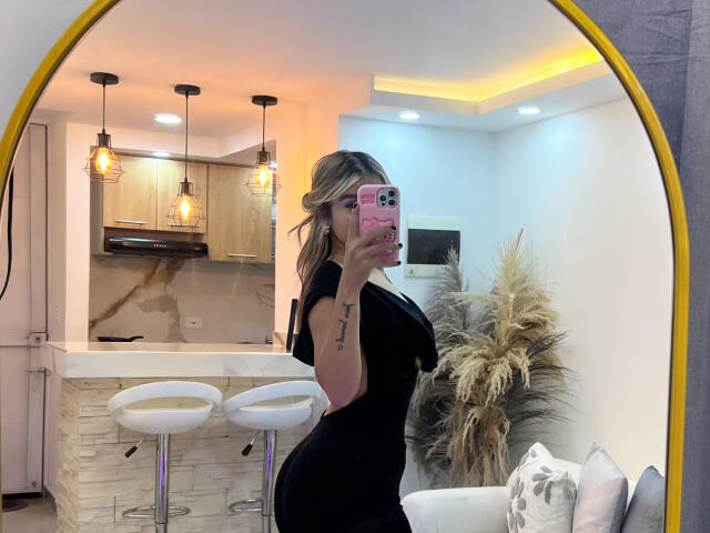 SashaPolet, con cabello rubio y un vestido negro, mostrando su gran trasero y pequeños senos en un selfie frente a un espejo, sosteniendo un teléfono rosa, en una cocina moderna..
