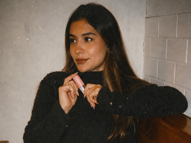Thalia Ares, con cabello largo y castaño, un suéter negro y peludo, sosteniendo un lápiz labial rosa, con una ligera sonrisa, ojos marrones, perforación en la nariz, en interiores, foto con textura, parte superior del cuerpo, mirando a la cámara..
