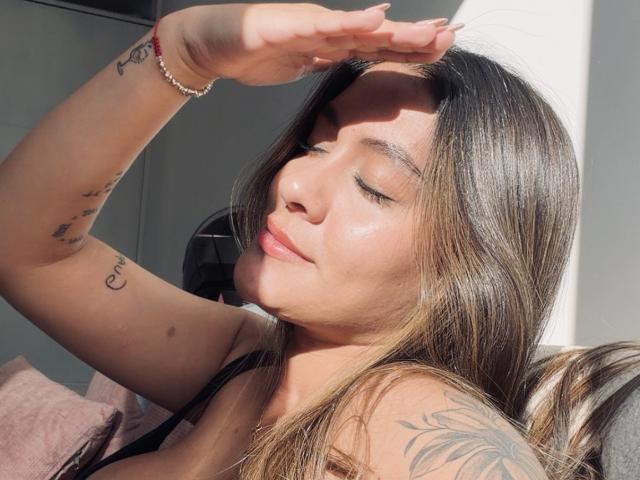 ThaliaAres, les yeux fermés, visage illuminé par le soleil, cheveux bruns, bras levé, tatouages sur le bras et l'épaule, sourire discret, peau rosée, bracelet, la lumière du soleil mettant en valeur sa peau..