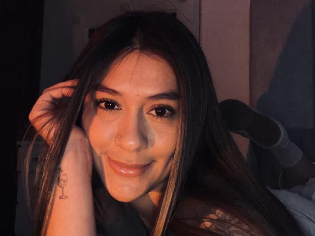 Thalia Ares, con el pelo largo y castaño, sonriendo, tiene un piercing en la nariz, un tatuaje en la muñeca y un líquido blanco en la barbilla, mirando a la cámara en una luz tenue..