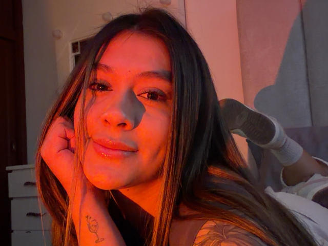 ThaliaAres com cabelos castanhos longos, luz avermelhada-alaranjada no rosto, sorriso discreto, tatuagem no pulso, deitada na cama, com o pé levantado, usando meias, iluminação suave, expressão relaxada..