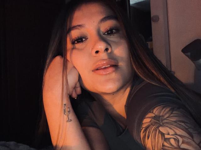 ThaliaAres, avec ses lèvres brillantes, ses longs cheveux bruns et un tatouage à son poignet droit, appuyée sur sa main, portant une chemise sombre, montrant un tatouage sur son bras, dans une lumière tamisée..