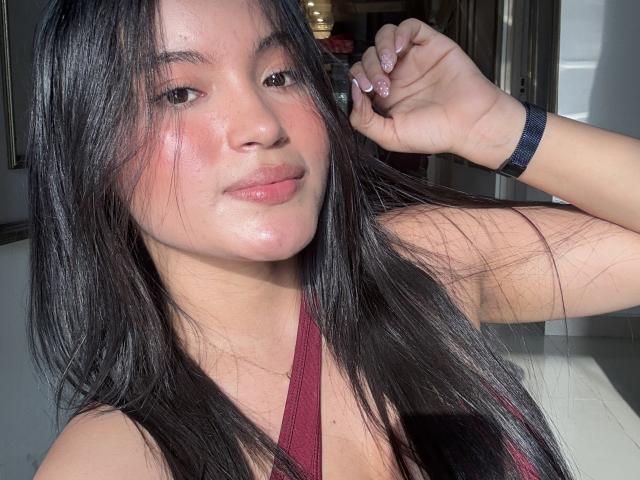 MiiaPalmer with long black hair, pink blush on cheeks, subtle smile, wearing a red halter top, pink nails, and a black wristband, sunlight on her face.