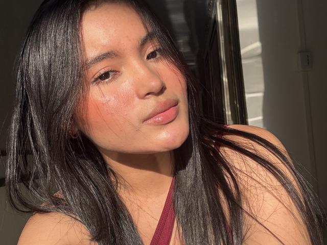 MiiaPalmer has long black hair, tan skin, and a cute face with pinkish cheeks, wearing a red halter top, sunlight highlighting her smooth skin and pouty lips.
