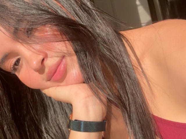 MiiaPalmer's face with long black hair, pink lips, and a subtle smile, resting her chin on her hand, wearing a green wristband, sunlight on her cheek.