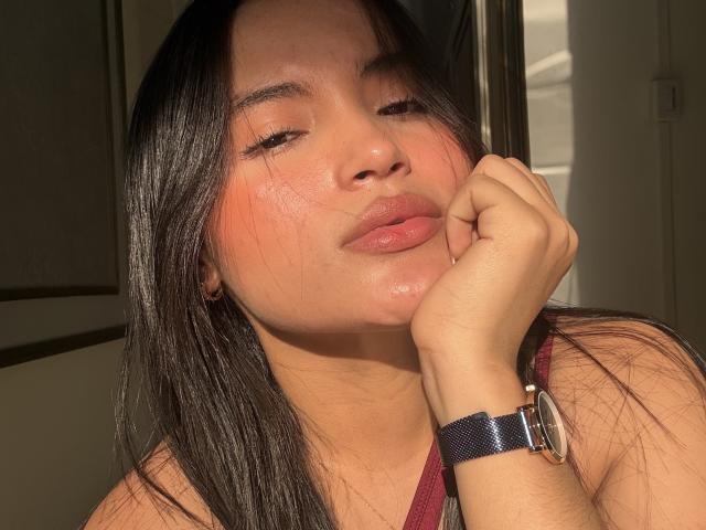 MiiaPalmer with dark straight hair, closed eyes, and slightly parted pink glossy lips, resting her chin on her hand, wearing a maroon top and a blue wristwatch.