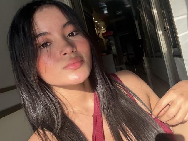 MiiaPalmer with long black hair, pinkish cheeks, and a slight smile, wearing a red top, sunlight highlighting her face, holding a lock of her hair.