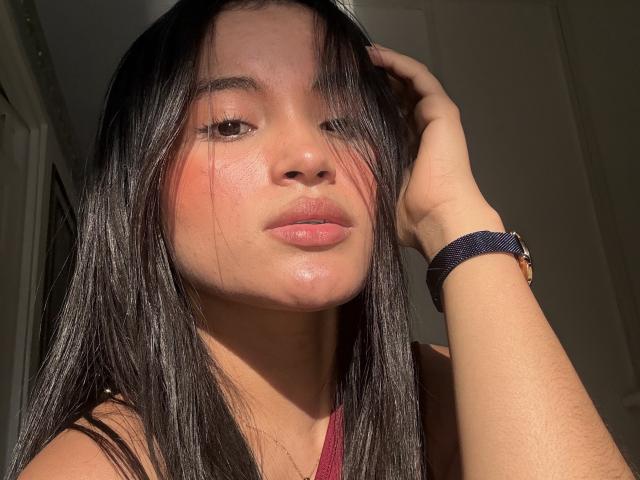 MiiaPalmer has shiny black hair, pinkish-red cheeks, and glossy lips. She wears a blue wristband, her hand in her hair, sunlight on her face. She looks sexy.