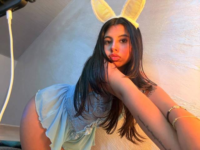 Celeste Morel, avec ses longs cheveux foncés, portant une robe blanche en dentelle, des oreilles de lapin jaunes et un bracelet, penchée en avant, faisant la moue avec sa main près de sa bouche, montrant un tatouage..