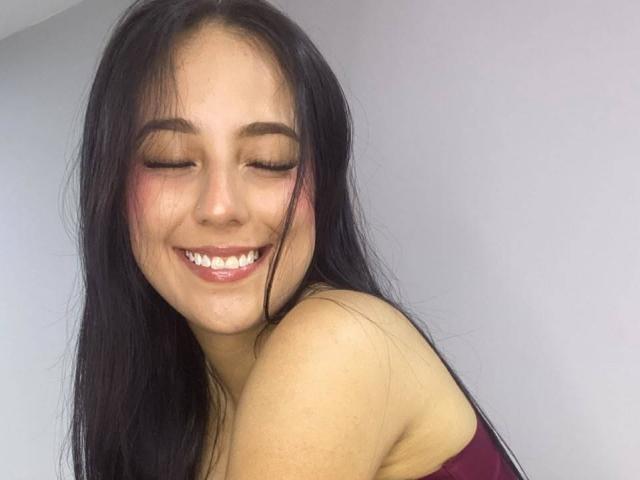 StefanyDelly has long black hair, closed eyes, and a big smile showing her white teeth. She's wearing a maroon top, and her ass is slightly visible.