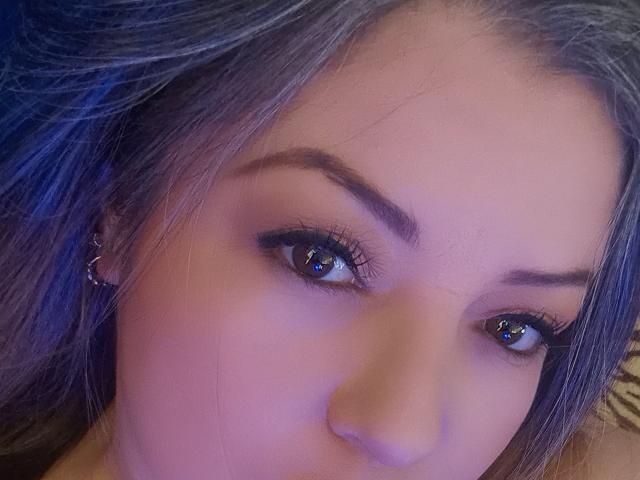 Le visage de NatalyHami, avec ses yeux bleus, ses longs cils et son maquillage discret. Elle a les cheveux gris-bleus et porte un petit anneau à son oreille gauche. Sa peau est claire..