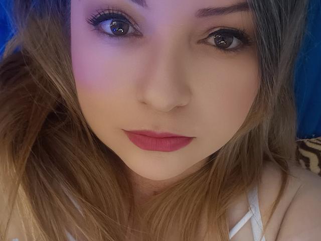 Il viso di NatalyHami, con i suoi adorabili occhi marroni, il rossetto rosa e i capelli castano chiaro, mostra una leggera sfumatura blu e viola sulla pelle. Il seno è parzialmente visibile sotto la maglietta bianca..