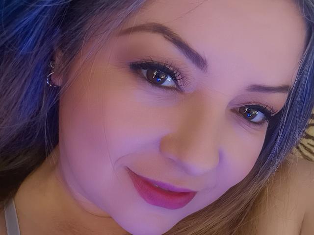 Het gezicht van NatalyHami, met blauwe ogen, paarse lippenstift en lichtbruin haar, draagt een zilveren oorbel, glimlacht zachtjes, toont een lichte grijns en haar ogen reflecteren licht..