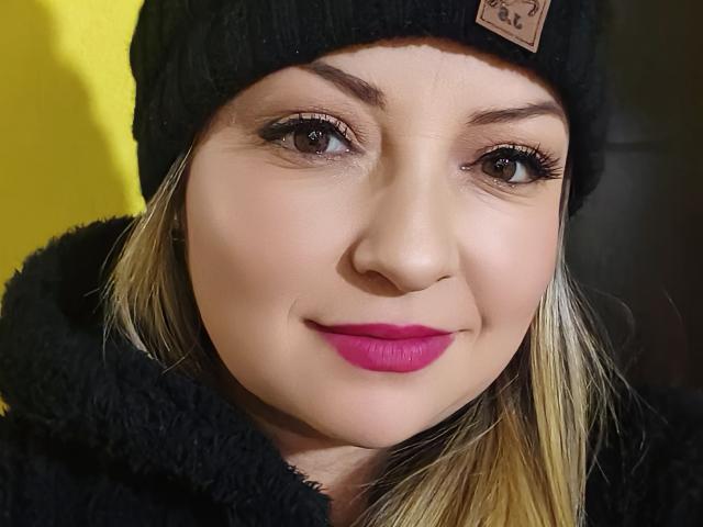 NatalyHami, avec ses cheveux blonds, son bonnet noir, son rouge à lèvres rose et son manteau noir duveteux, sourit avec ses yeux marrons et un maquillage discret. Son visage est centré, et elle a l'air adorable..
