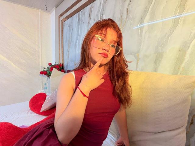 Elodie Gardner, con i capelli rossi, gli occhiali e un vestito rosso, che si morde un dito, seduta su un divano bianco con un cuscino a forma di cuore rosso e un vaso di fiori sullo sfondo..
