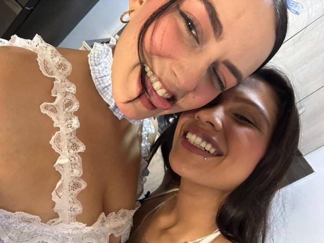 ElizabethAndEliana with their tongues out, smiling at the camera. ElizabethAndEliana has white lace boobs, a nose ring, and dark hair. Eliana has a nose piercing and brown hair.