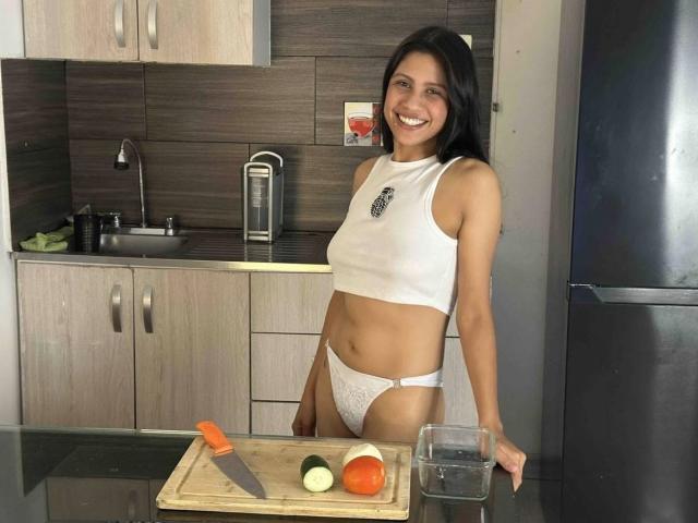 ElizabethAndEliana with dark hair, smiling, wearing a white crop top and white lace panties, standing in a kitchen with a wooden cutting board, knife, and veggies.