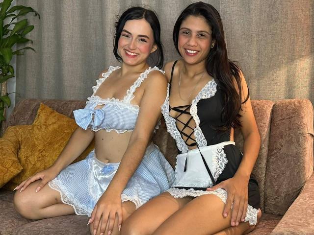 ElizabethAndEliana with dark hair, smiling, wearing blue and black lingerie with lace, sitting on a couch, showing their tits and small panties, hands on their thighs.