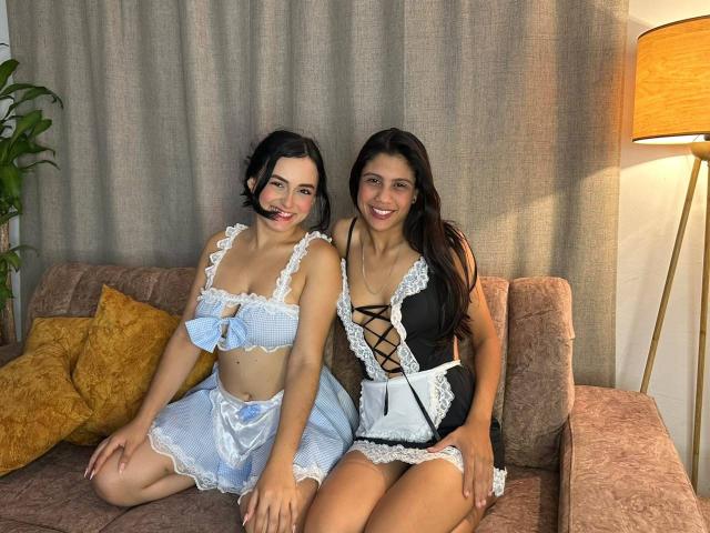 ElizabethAndEliana with dark hair, wearing blue and white maid outfits, sitting on a pink couch. ElizabethAndEliana has small tits, blue bow, smiling. Eliana has black lingerie, smiling.