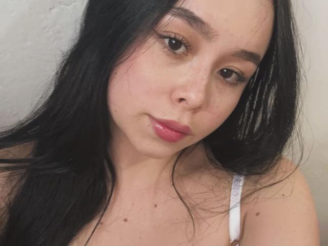 NikiSweety69 tem cabelos pretos longos, olhos castanhos, batom rosa e um leve sorriso, vestindo uma alça branca. Seus seios estão visíveis, com algumas pequenas manchas na região do peito..