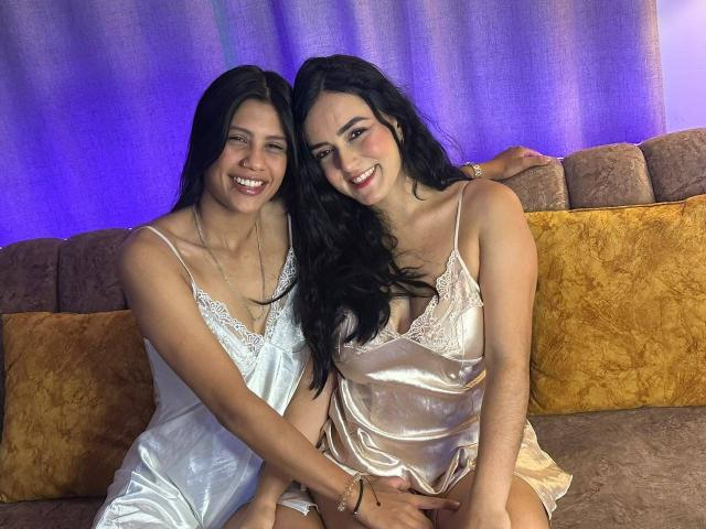 ElizabethAndEliana with long black hair, smiling, wearing silky white and pink nighties, sitting on a brown couch, hands on each other's laps, blue background. Boobs, cute, sexy.