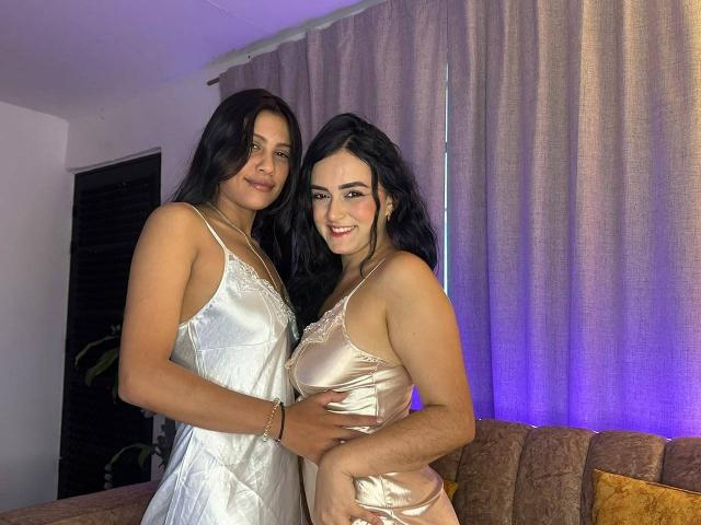 ElizabethAndEliana with dark hair, in silky white and gold dresses, standing in a room, smiling, Elizabeth's hand on Eliana's ass, both showing breasts, Eliana's hand on Elizabeth's hip.