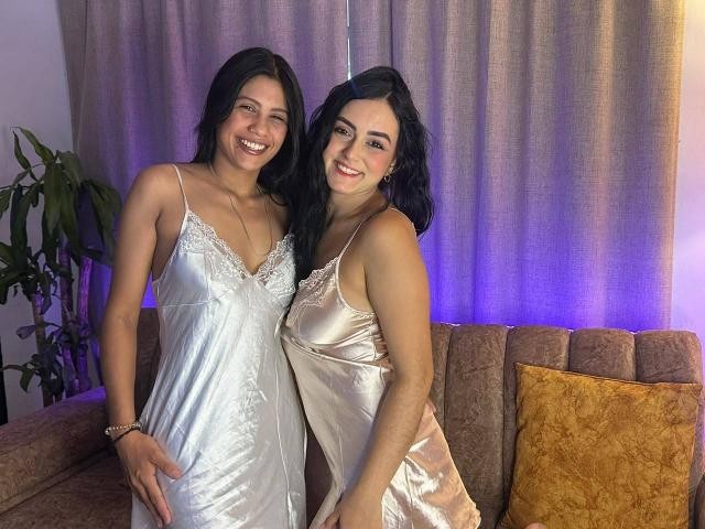 ElizabethAndEliana with black hair, smiling, wearing silky white and beige nightgowns, standing on a brown couch, both showing their tits, hands on their asses, soft purple lighting.