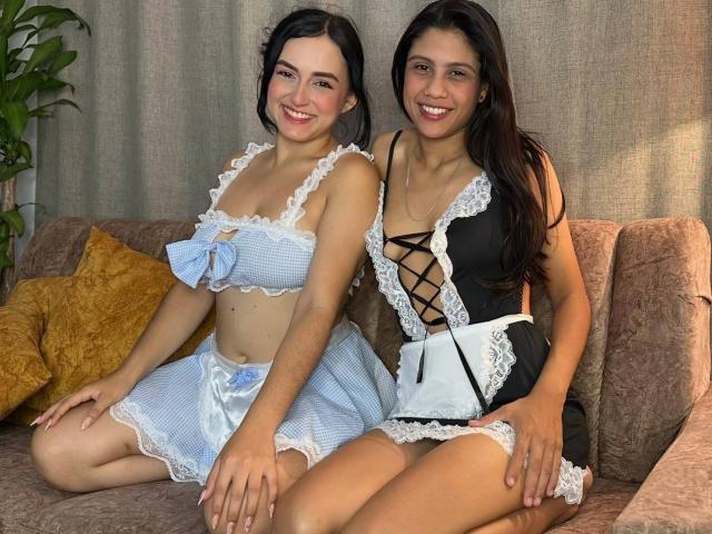 ElizabethAndEliana, two cute girls in French maid outfits, sitting on a couch. Elizabeth has small tits, light skin, and dark hair; Eliana has medium tits, tan skin, and black hair.