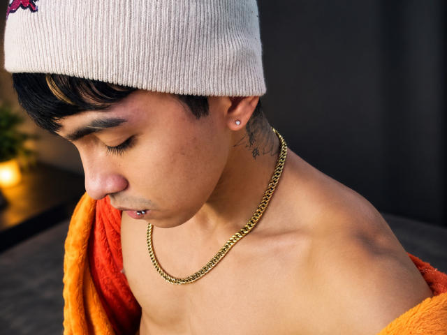 DrakeJack, con un berretto, una catena d'oro e un asciugamano arancione, ha un orecchio perforato, capelli neri corti e un tatuaggio, guarda in basso con un leggero broncio, senza maglietta, con la pelle abbronzata..