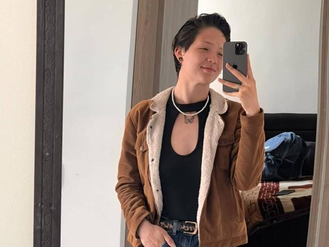 LissyLuxe with short black hair, smiling, wearing a brown velvet jacket with white fur, black tank top, and blue jeans, taking a mirror selfie with a smartphone.
