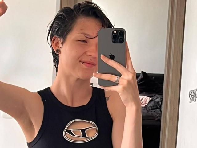 LissyLuxe with short wet black hair, sticking out tongue, holding iPhone, wearing black sleeveless shirt with oval cutout, showing small bare breast, mirror selfie, wearing black ear gauges.